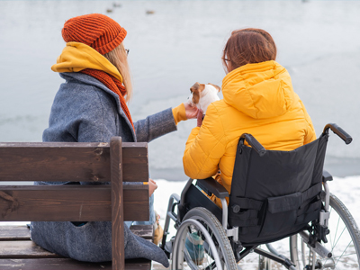 Zwei Frauen sitzen am Ufer eines Gewässers. Eine Frau in einem Rollstuhl trägt eine gelbe Jacke und füttert einen kleinen Hund, während die andere Frau in einem grauen Mantel und orangenem Hut neben ihr sitzt.